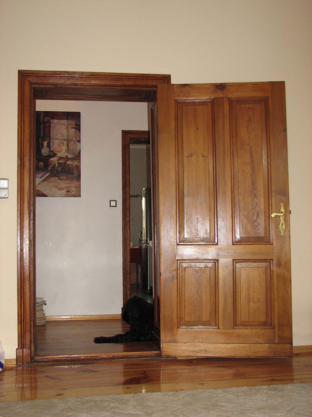 Otwarta, drewniana, czteropanelowa brama z mosiężną klamką, widoczna z perspektywy niskiego kąta, z fragmentem podłogi i czarnym psem leżącym w przejściu.